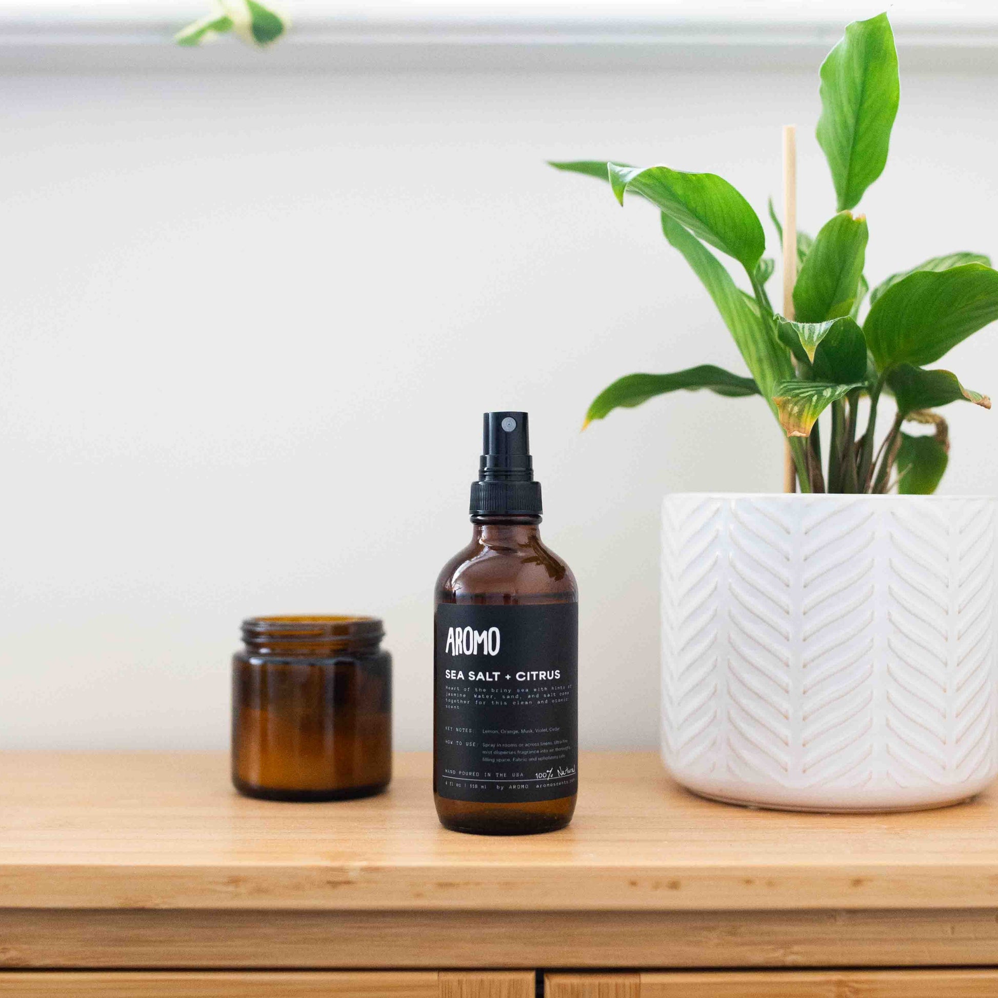 full sized aromo sea salt citrus room and linen spray atop a bathroom cabinet accompanied by an amber glass candle and a small houseplant
